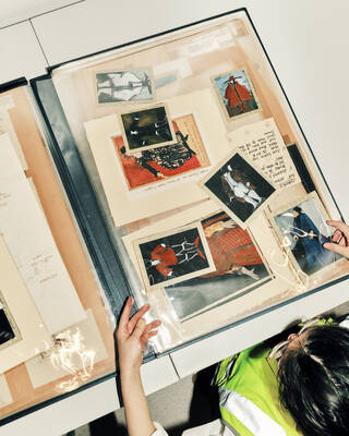 A large scrap book is open on a white desk. there are black and white photos, overlapping other pieces of paper with images and texts. The pages look as if they are covered with plastic